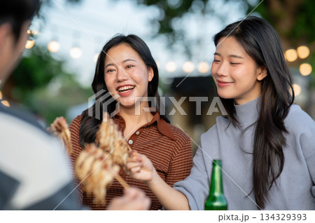 Asian women and a man friend eating meat skewers while sitting at table in a campground restaurant. 134329393
