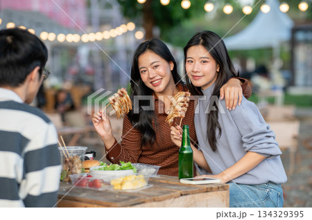 Asian woman eating meat skewer and hugging friend while sitting at table in a campground restaurant. 134329395