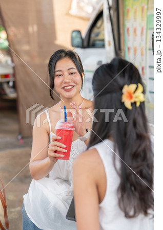 Pretty asian woman holding drinking juice and talking with her friend while sitting aside food truck 134329997