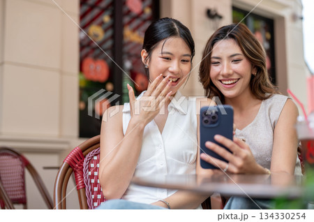 Pretty asian woman showing phone to a friend covering smiling mouth while sits at table outside cafe. Pretty asian woman showing phone to a friend covering smiling mouth while sits at table outside cafe. 134330254