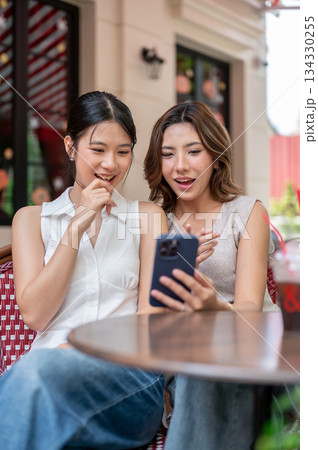 Pretty asian woman showing phone to a friend with hand holding chin while sits at table outside cafe Pretty asian woman showing phone to a friend with hand holding chin while sits at table outside cafe 134330255
