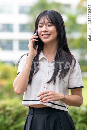 Pretty asian woman talking on smartphone with a smile while standing in green garden cafe or a park. 134330367