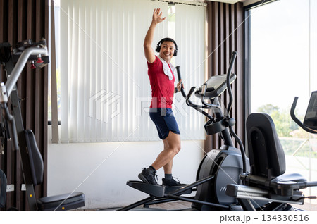 Full body shot of healthy man wearing headphones waving a hand while using exercise equipment in gym Full body shot of healthy man wearing headphones waving a hand while using exercise equipment in gym 134330516