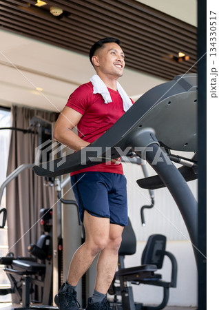 Smiling asian middle aged healthy man with towel on shoulder running exercising on treadmill in gym. Smiling asian middle aged healthy man with towel on shoulder running exercising on treadmill in gym. 134330517