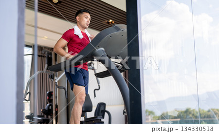 Asian middle aged strong healthy man with towel on shoulder running or exercising on treadmill in gym Asian middle aged strong healthy man with towel on shoulder running or exercising on treadmill in gym 134330518