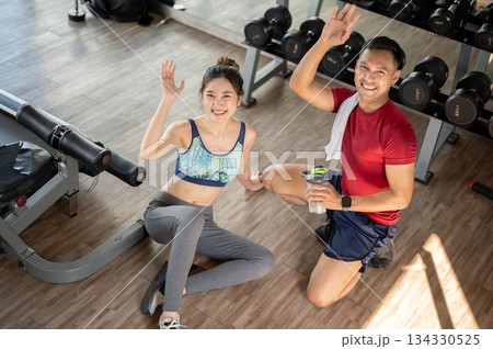 Pretty woman client and fitness trainer man waving hand as sitting on floor after exercising in gym Pretty woman client and fitness trainer man waving hand as sitting on floor after exercising in gym 134330525