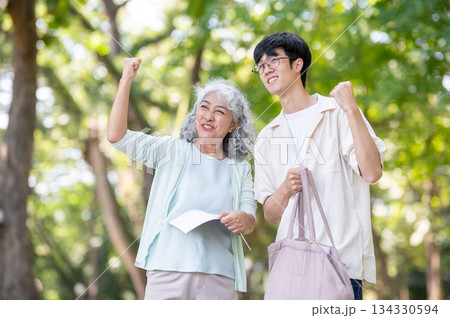 Old asian woman grandmother holding book and young man grandson making a fist while walking in park. 134330594