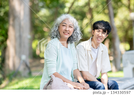 Old asian woman grandmother smiling while sitting on bench with a young man grandson in the park. 134330597