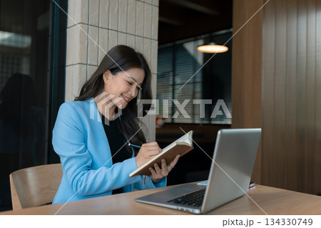 Female hand taking notes on a notebook handwritten note writing business schedule and use a laptop computer at the office desk in the morning. 134330749