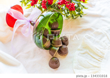 Chocolates are poured out of a green glass glass onto a white cloth with festive decor in the background.  134331018