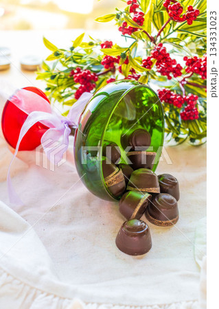 Chocolates are poured out of a green glass glass onto a white cloth with festive decor in the background. 134331023