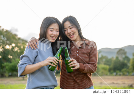 Asian woman friends standing and clinking drink bottles in a grass field with mountain view and sky. Asian woman friends standing and clinking drink bottles in a grass field with mountain view and sky. 134331894