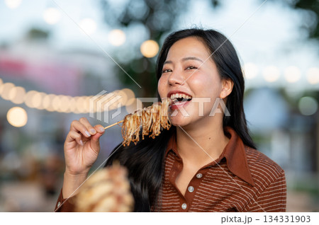 Pretty asian woman eating a delicious meat skewer while sitting at table in a campground restaurant. 134331903