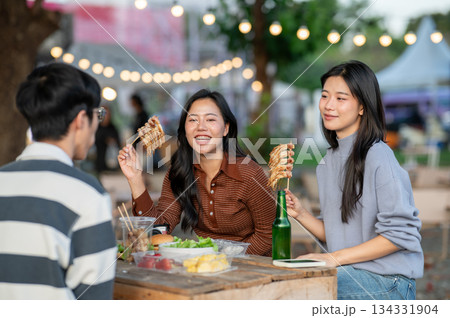 Asian women eating meat skewers and talking to man friend sitting at table in campground restaurant. Asian women eating meat skewers and talking to man friend sitting at table in campground restaurant. 134331904
