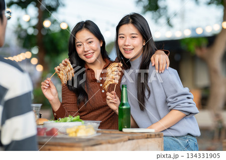 Asian woman eating meat skewer and hugging a friend while sitting at table in campground restaurant. Asian woman eating meat skewer and hugging a friend while sitting at table in campground restaurant. 134331905
