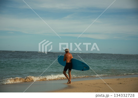 Man strolls on the beach holding a surfboard near the waters edge 134332728