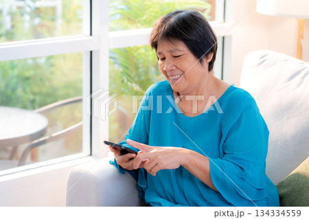 Senior asian woman smile while using on smartphone sitting on sofa in living room at home. 134334559