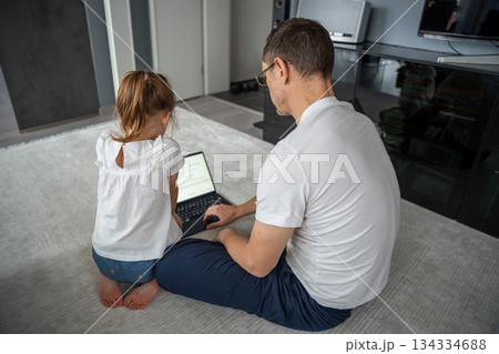 Father and daughter sitting on the carpet and using a laptop together. Digital home education and shared screen activities. 134334688