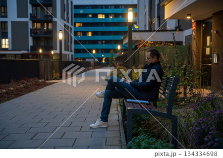 Man sitting on a bench in a modern residential courtyard in the evening. Urban lifestyle and moments of relaxation. 134334698
