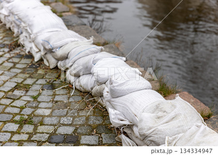 Sandbags form protective barrier riverbank reinforcing stone embankment against flood erosion. Urban waterfront shows emergency preparedness infrastructure wet surfaces reflections overcast weather 134334947