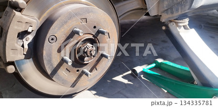 A close-up of the rear brake system of a dirty car with the wheel removed, showing the shiny brake rotor and caliper, and a green jack underneath. A close-up of the rear brake system of a dirty car with the wheel removed, showing the shiny brake rotor and caliper, and a green jack underneath. 134335781