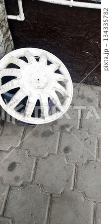 A dirty, white plastic wheel cover lies on a grey interlocking paved surface, resting against a dark wooden background and white pipes A dirty, white plastic wheel cover lies on a grey interlocking paved surface, resting against a dark wooden background and white pipes 134335782