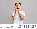 Surprised boy with hands on face looking at camera, emotional shocked child expressing amazement and astonishment on gray background, studio portrait of kid reacting with wide eyes and open mouth 134335827