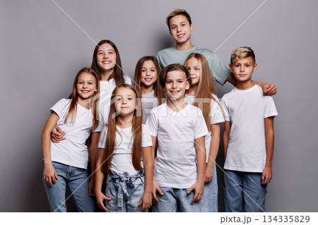 Group of happy children, boys and girls, standing together and smiling in studio against gray background. Beautiful kids wearing white t-shirts showing friendship, joy and positive emotions 134335829