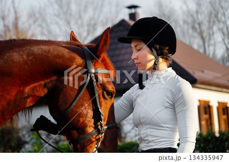 young Caucasian woman stands next to a horse, looking at it, wearing a riding outfit. A professional show jumper stands next to the horse, holding its bridle young Caucasian woman stands next to a horse, looking at it, wearing a riding outfit. A professional show jumper stands next to the horse, holding its bridle 134335947