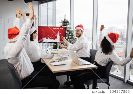 Successful team of multiethnic business people in Santa hats raising hands and screaming with happiness while working together in decorated with Christmas tree office in business center. Successful team of multiethnic business people in Santa hats raising hands and screaming with happiness while working together in decorated with Christmas tree office in business center. 134337896