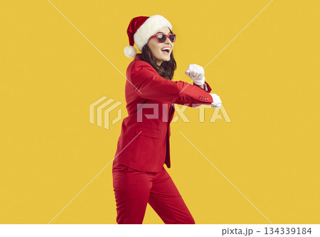 Joyful young woman in red suit, sunglasses and Christmas hat dancing on yellow background 134339184