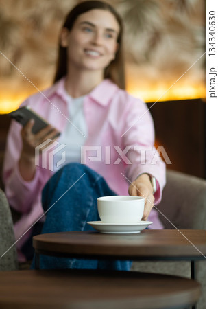 Cute young woman in pink with a phone in hands having tea Cute young woman in pink with a phone in hands having tea 134340630