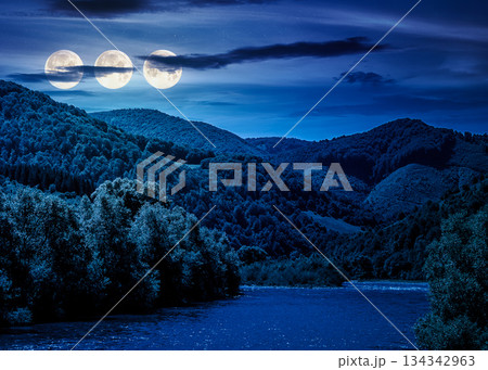 river flows through green forest near mountain at night. picturesque nature of rural area on serene summer weather under clouds on sky in full moon light. backdrop for fiction art or mystery concepts 134342963
