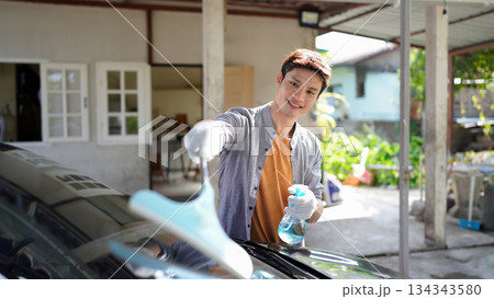 A man washing a car with a spray bottle and cloth outdoors, smiling while cleaning with an energetic and positive mood 134343580