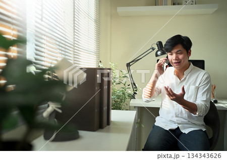 A man talking on the phone in a bright home office, expressing ideas with natural gestures in a relaxed work atmosphere. A man talking on the phone in a bright home office, expressing ideas with natural gestures in a relaxed work atmosphere. 134343626