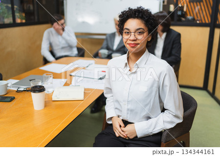 Portrait of young female team leader businesswoman in office, colleagues meeting in background. Confident African American woman business leader sitting in office. Boss leader professional worker 134344161