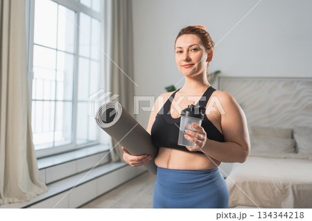 Yoga fitness sport workout training. Woman hold yoga mat bottle water ready for practicing yoga at home. Plus size female doing sports indoor. Girl in sportswear have training workout for weight loss Yoga fitness sport workout training. Woman hold yoga mat bottle water ready for practicing yoga at home. Plus size female doing sports indoor. Girl in sportswear have training workout for weight loss 134344218