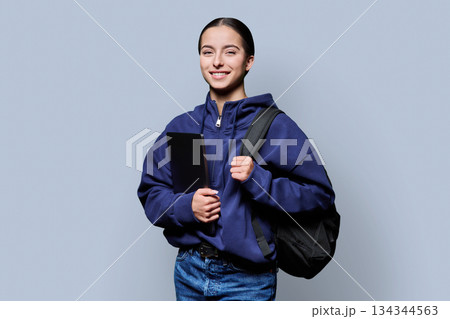 Portrait of beautiful female teenage student with backpack, laptop on gray background 134344563