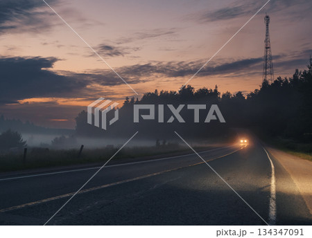 A vehicle with headlights on drives down a dark asphalt road, in a rural area near a forest A vehicle with headlights on drives down a dark asphalt road, in a rural area near a forest 134347091
