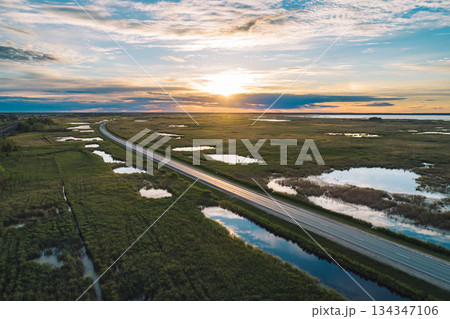 Aerial view of long rural road crossing through marshland at golden hour sunset 134347106