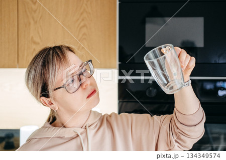 A woman wearing glasses and a hoodie examines a clear glass cup in her kitchen. Kitchen cleanliness, domestic hygiene, household routine, attention to detail. A woman wearing glasses and a hoodie examines a clear glass cup in her kitchen. Kitchen cleanliness, domestic hygiene, household routine, attention to detail. 134349574