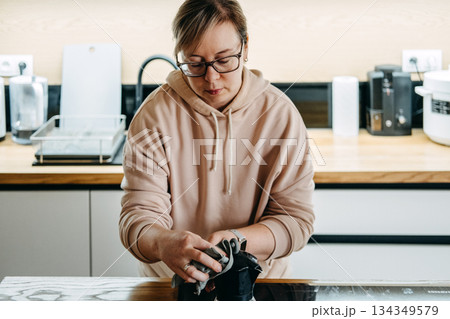 A woman wearing glasses and a hoodie wipes a black kitchen appliance on the counter with a dark reusable cloth. Eco friendly kitchen, reusable cloths, sustainable cleaning products, domestic hygiene. 134349579