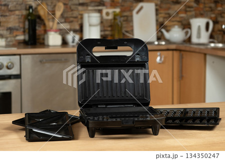 Open Electric Sandwich Maker and Waffle Grill with Interchangeable Plates on a Kitchen Table. 134350247