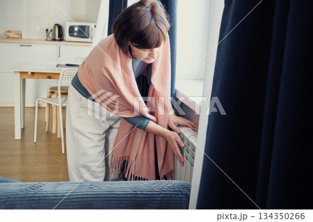 Senior woman feeling the warmth of radiator, coping with rising heating costs 134350266