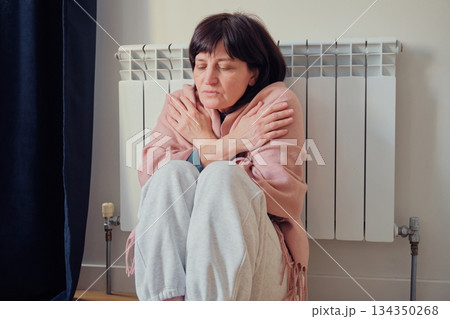Woman shivering and hugging herself while sitting next to non-working radiator, feeling cold indoors 134350268