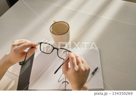 Hands holding eyeglasses over notebook and pen on table with morning sunlight 134350269