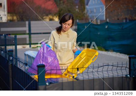 Woman wearing gloves placing garbage bag into outdoor recycling container 134351209