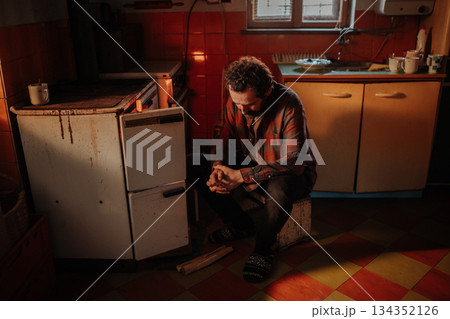 Despaired man sitting by wood stove in aged interior. 134352126