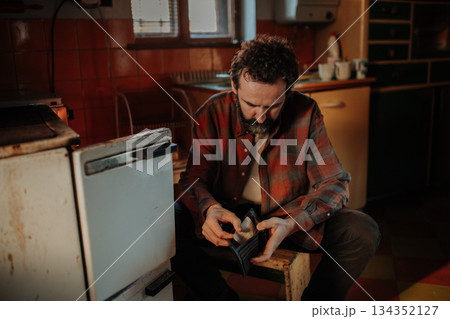 Despaired man sitting by wood stove looking into empty wallet. 134352127