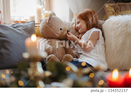 Small girl sitting on sofa indoors at home at Christmas, hugging teddy bear. Small girl sitting on sofa indoors at home at Christmas, hugging teddy bear. 134352157
